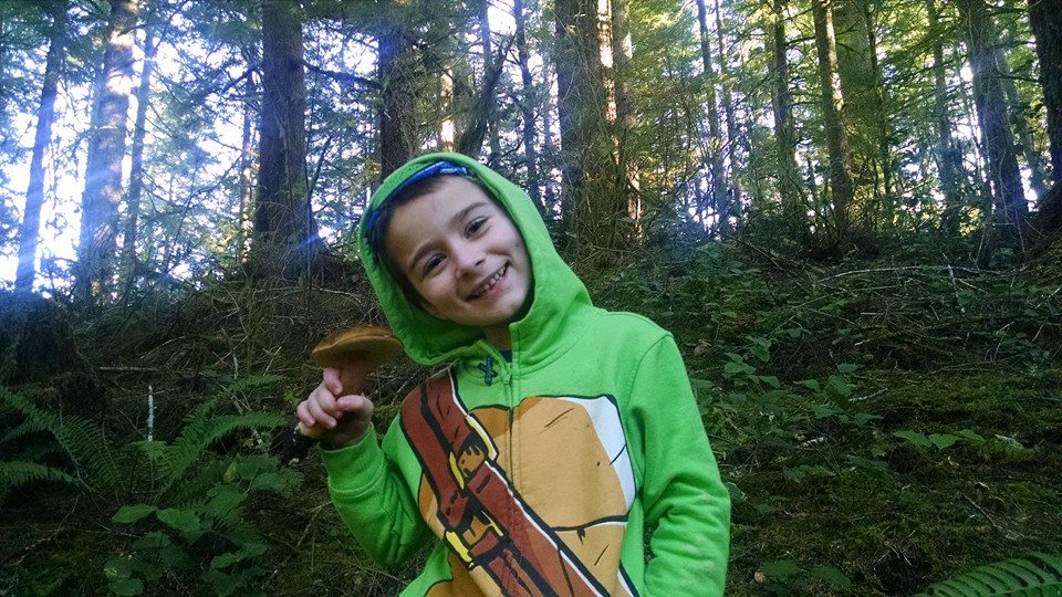 A kid smiling in the forest, holding up a mushroom find